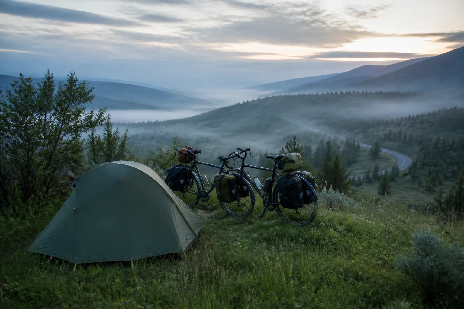 Small touring camp at dawn with two bicycles beside a tent, mist over a looping valley road in the distance, cool blue light, 1200x800